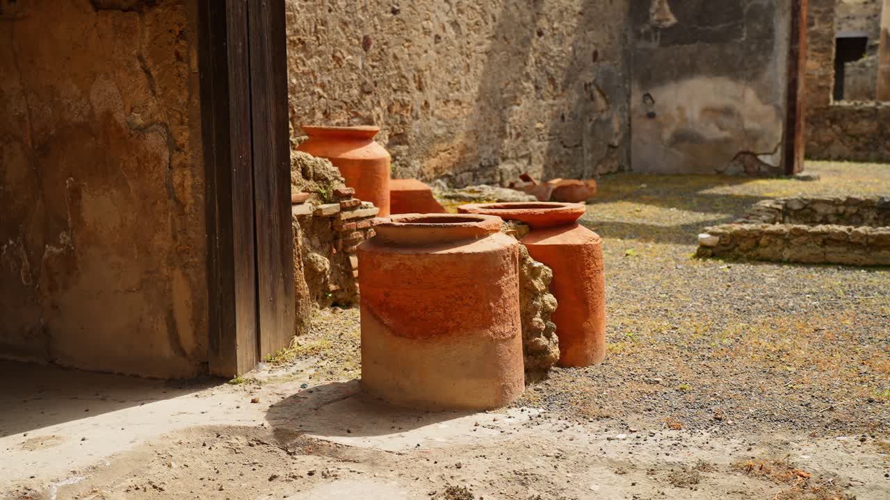 Movimento cinematografico oltre una porta verso giare di terracotta a Pompei, che cattura vasi di stoccaggio romani illuminati dal sole e rovine di cortili consumate dal tempo con una calda atmosfera archeologica.