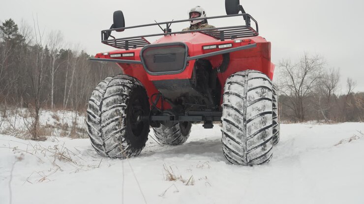 Pilota caucasico che corre con un ATV rosso attraverso un campo innevato, ampio paesaggio con sfondo di foresta di pini, tracce di pneumatici visibili e accelerazione, scatti frontali dinamici che mostrano movimento e controllo, inverno energico