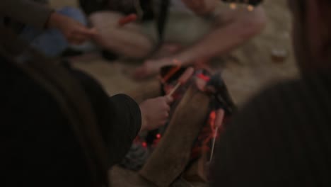 grupo de jóvenes amigos sentados junto al fuego en la playa, asando salchichas y tocando la guitarra 1