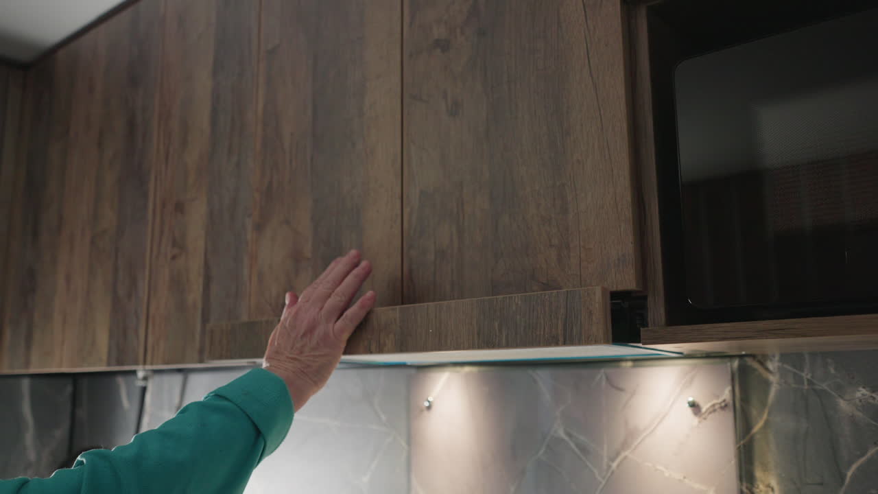 Older woman opening kitchen cabinet door in modern kitchen, focused on retrieving something from upper shelf, with kitchen tools, countertops, and appliances visible in background