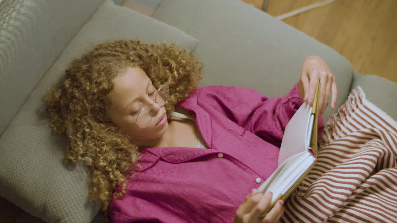 Cute blonde girl lying on couch reading book, turning pages