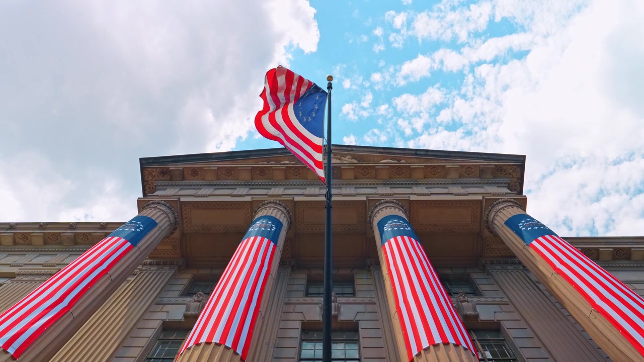 Vista em ângulo baixo de faixas patrióticas em um edifício governamental, Washington D.C. Colunas envoltas em tecido vermelho, branco e azul com a bandeira tremulando contra o céu nublado.