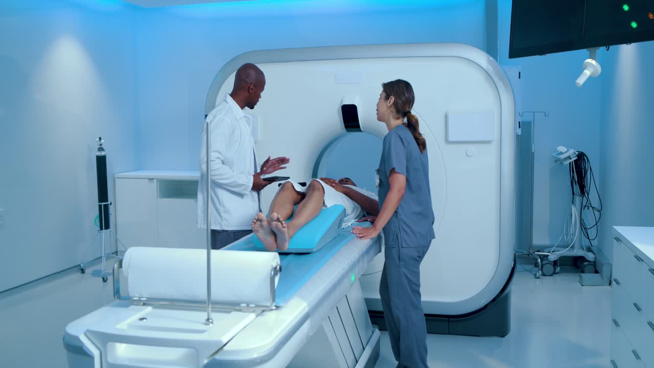 Paziente con tecnico e medico entrano da sinistra controllando il tablet, il personale regola un cuneo blu per la risonanza magnetica.