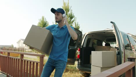 joyful deliveryman enjoyig music with headphones and carrying parcel to house from van