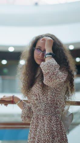 woman in vintage dress poses by iron railing in spacious mall, adjusting her hair with a thoughtful expression, background features blurred shoppers, soft lighting, and architectural details