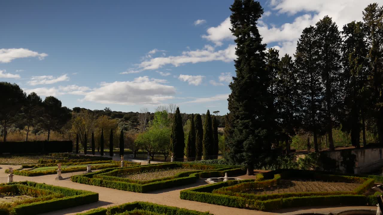 tiro estático de un jardín barroco con árboles un cielo azul con nubes moviéndose hacia la izquierda
