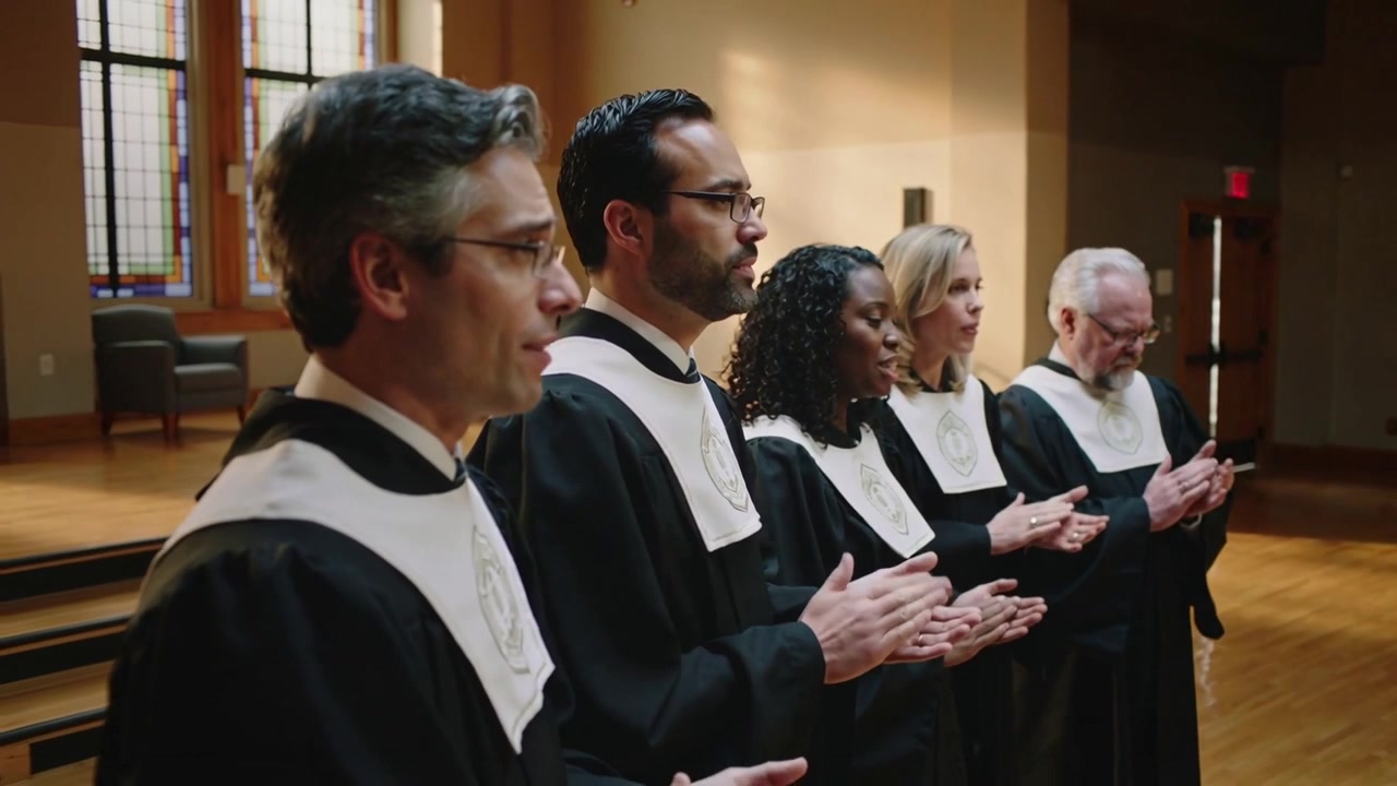 Inquadratura che si allarga su cinque coristi che cantano e applaudono in chiesa, mentre guidano la funzione religiosa indossando tuniche.
