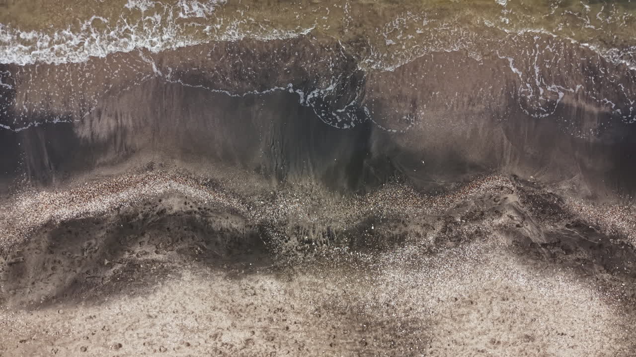 Le onde si infrangono su una spiaggia sabbiosa con una costa scura di giorno, in una zona costiera con ciottoli e conchiglie sparse