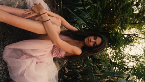 horizontal shot of full body portrait of gorgeous young brunette woman sitting on ground wearing pink gown in a garden