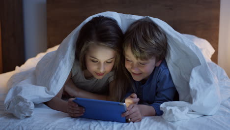 linda hermanita y hermano acostados debajo de la manta y viendo algo en la tableta por la noche