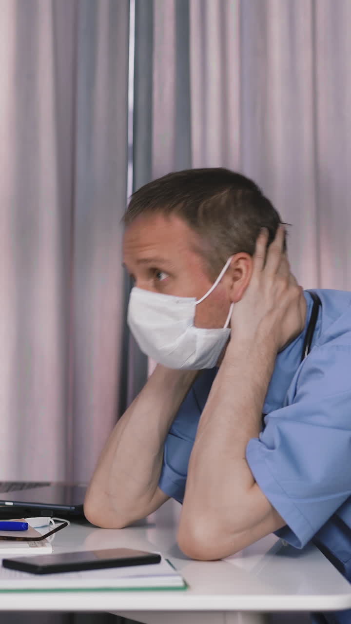 overstrained male doctor wearing blue uniform and protective mask sits at table with laptop and medical records in modern hospital office