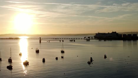 Boats-silhouetted-on-golden-shimmering-harbour-ocean-surface-aerial-rising-view