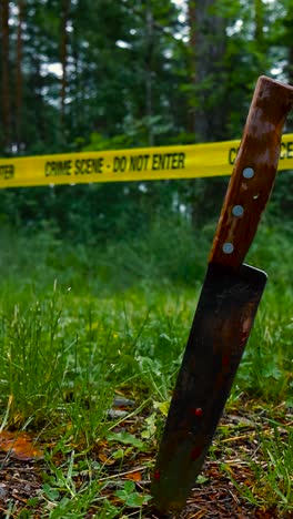bloody knife vertically standing in front of a yellow crime scene do not enter tape at a forest during a cloudy and wet day. the knife is stabbed in the grassy ground and background is blurry bokeh