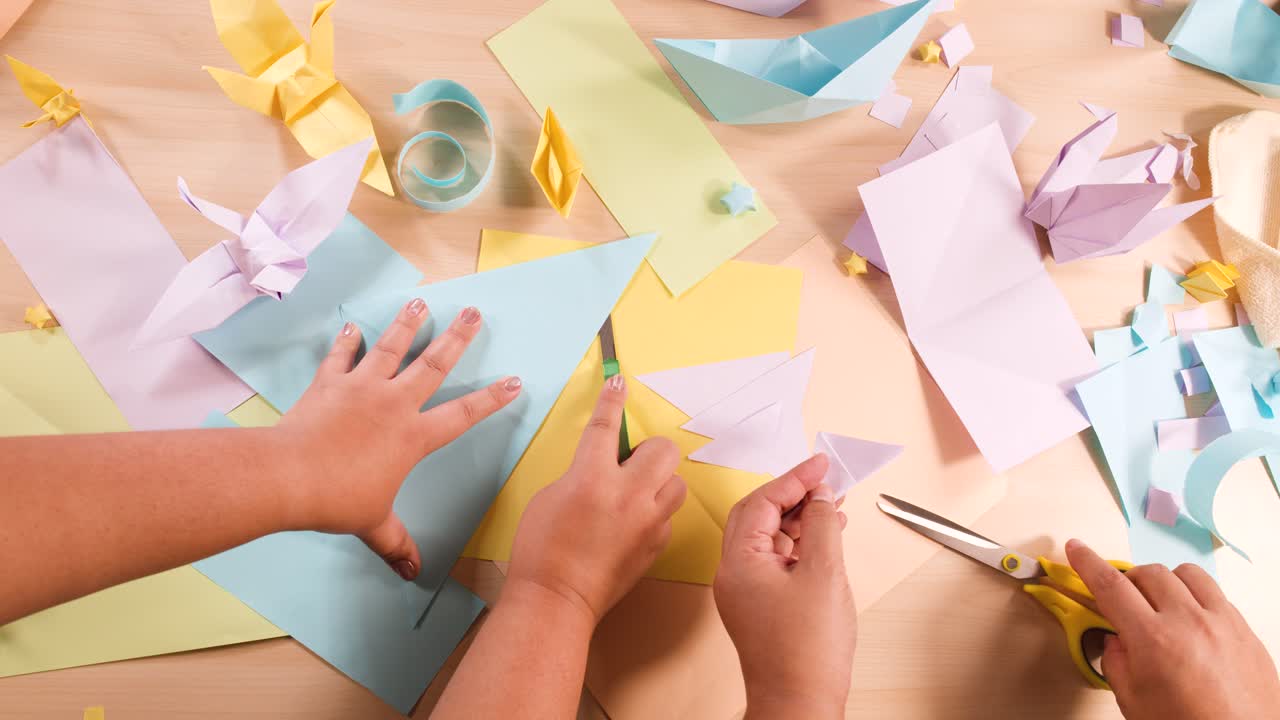 Top-down shot of hands meticulously folding and cutting pastel paper for various origami projects