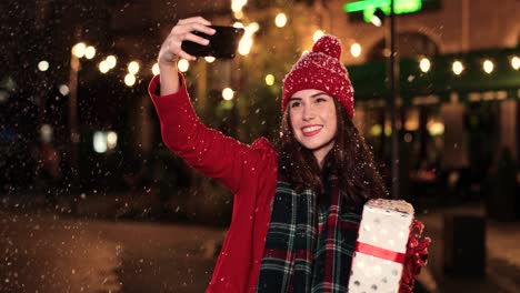 vista de cerca de una mujer caucásica con abrigo rojo sosteniendo un regalo y haciéndose un selfie con un smartphone en la calle mientras nieva en navidad