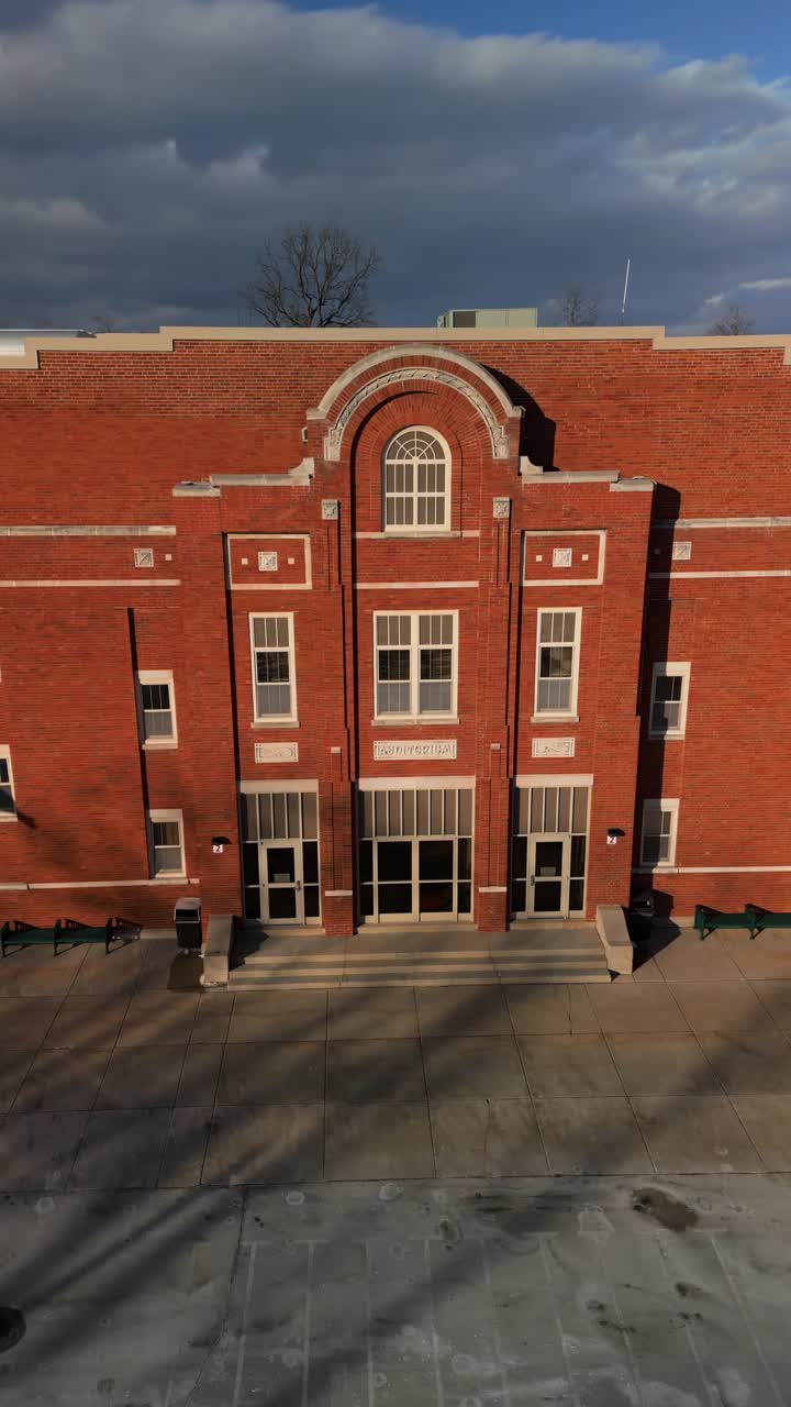 Suburban brick school under bright sky showing entry path, facade, and windows in sunlight, vertical aerial orbit