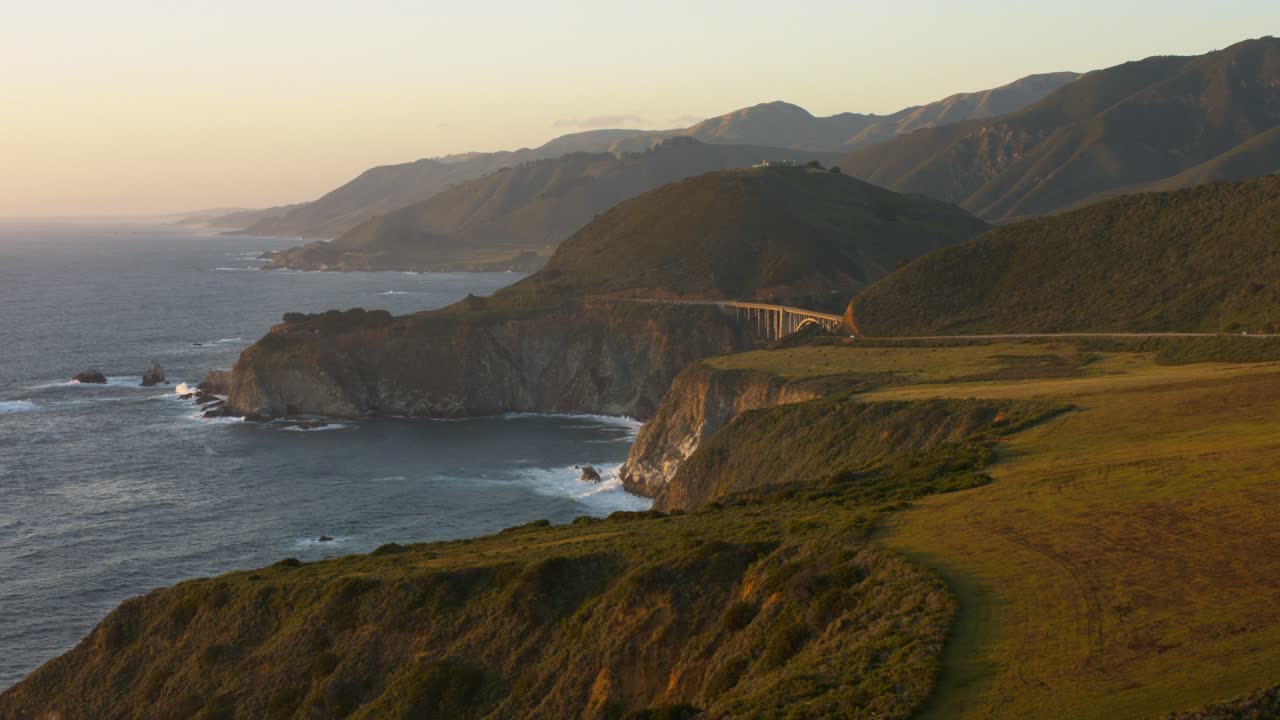 Stunning aerial view of Bixby Bridge during golden hour, showcasing the breathtaking Big Sur coastline with ocean waves, steep cliffs, and surrounding mountains in soft sunlight