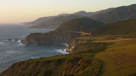 stunning aerial view of bixby bridge during golden hour, showcasing the breathtaking big sur coastline with ocean waves, steep cliffs, and surrounding mountains in soft sunlight