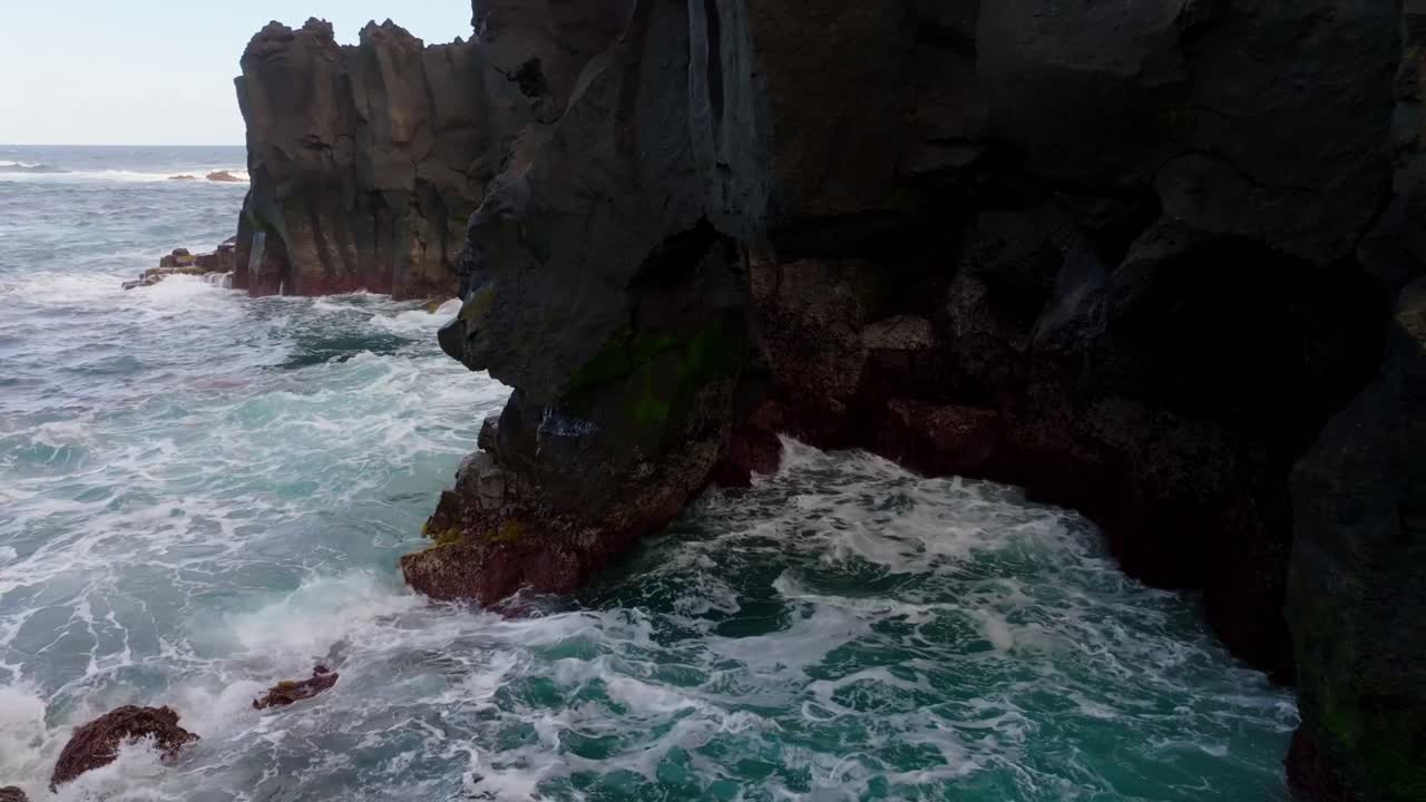 Aerial view of black volcanic rock formations meeting turquoise Atlantic waters, with a striking white building perched along the rugged Ferraria coast in the Azores