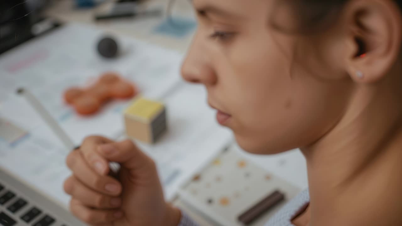 Donna concentrata in top casual che tiene una penna vicino alla bocca mentre studia e giocherella con il portatile sulla scrivania di casa, spazio per la copia
