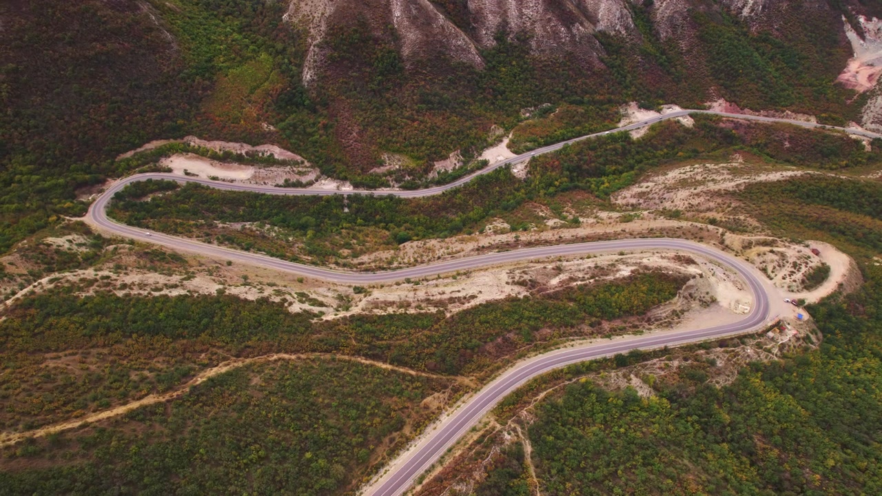 Vista aérea que muestra curvas cerradas conectadas serpenteando por laderas de montañas verdes y rocosas, mientras el dron oblicuo elevado revela la geometría de la carretera, la escala del terreno y el paisaje de viaje remoto en Daguestán