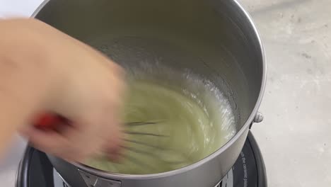 Close-up-shot-of-a-professional-pastry-chef-hand-mixing-and-whisking-egg-white-with-steaming-hot-water-on-electric-cooktop-in-a-commercial-bakery-setting