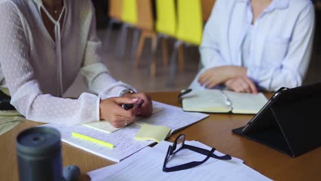 two attractive women smiling, having a conversation in a library with shelves of books on background. sitting at the wooden table with tablet and diverse papers on on it. students life, exams, preparation