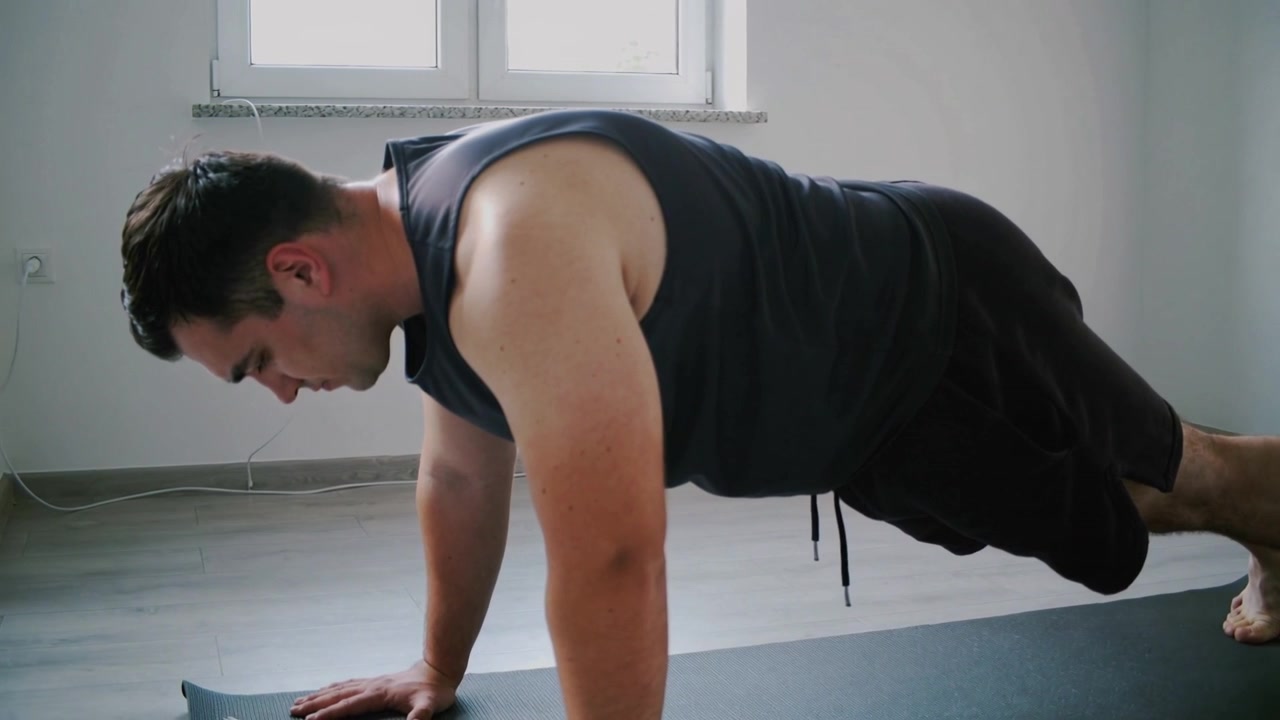 Uomo in allenamento che indossa canotta e pantaloncini, mantiene la posizione di plank alto in una stanza su un tappetino da esercizio vicino a un cavo della finestra