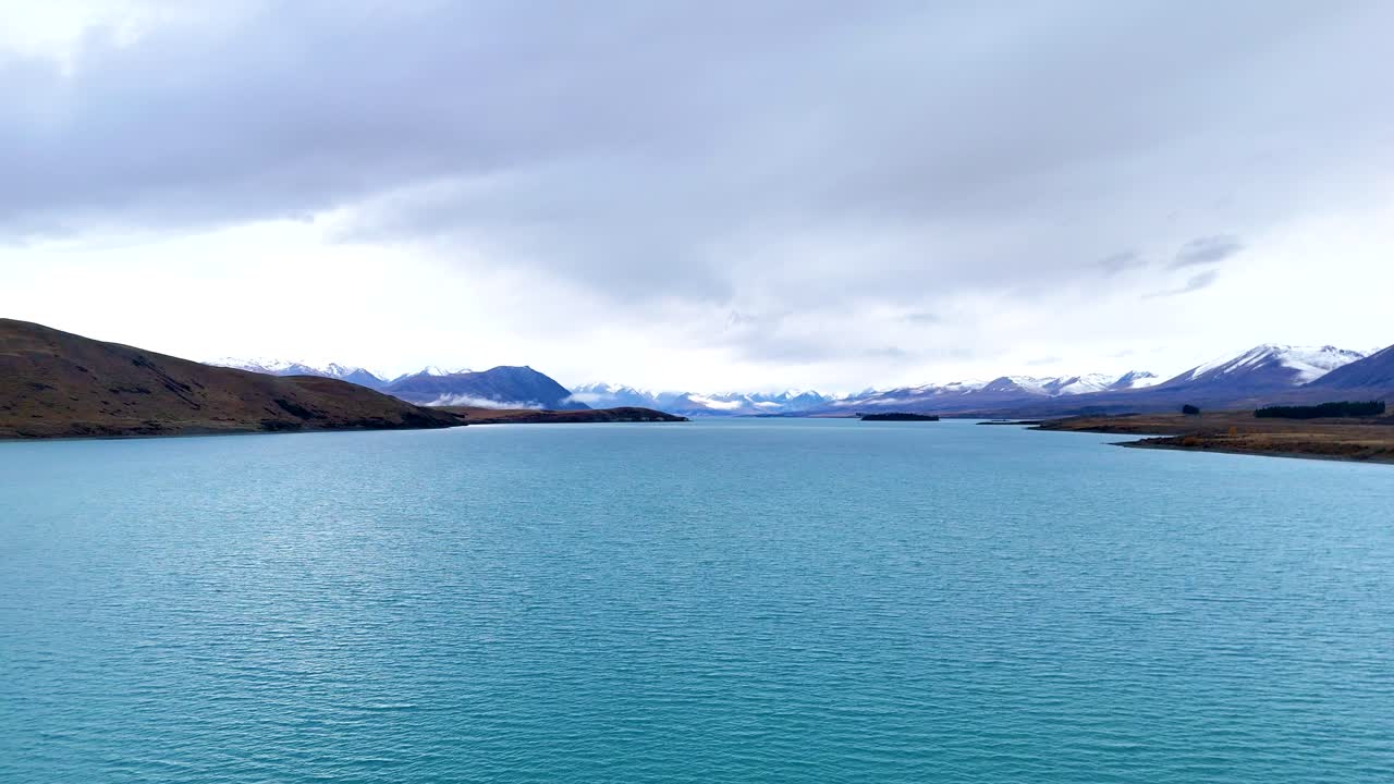Tranquil Lake Tekapo with turquoise waters and distant snow-capped mountains under overcast skies. Captured in Canterbury, New Zealand