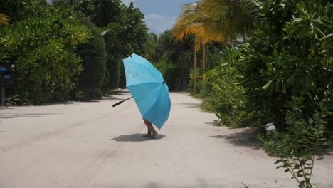 a cute small child hidden under a big blue umbrella walking barefoot on a sandy beach road with green palm trees on maldivian resort island on a clear summer day