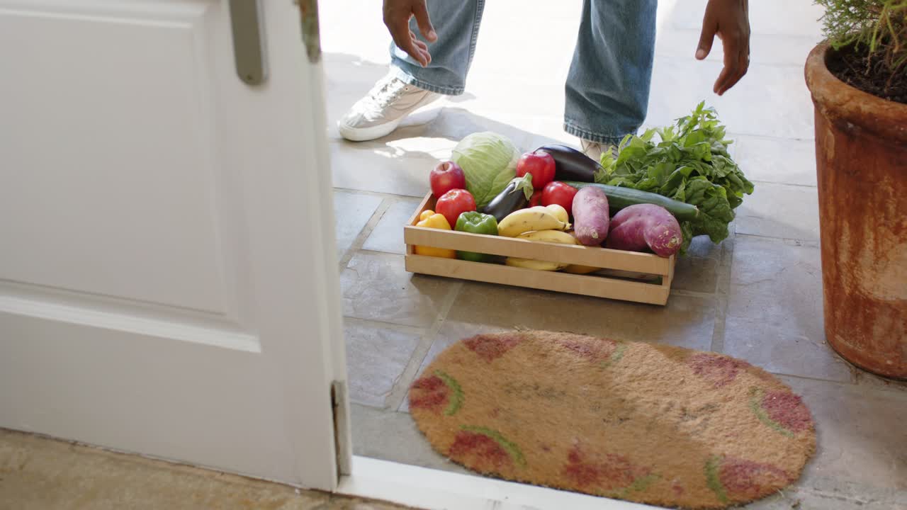Homem africano de jeans e tênis vendo uma caixa de hortifrúti na porta, se movendo e carregando-a para dentro de casa.