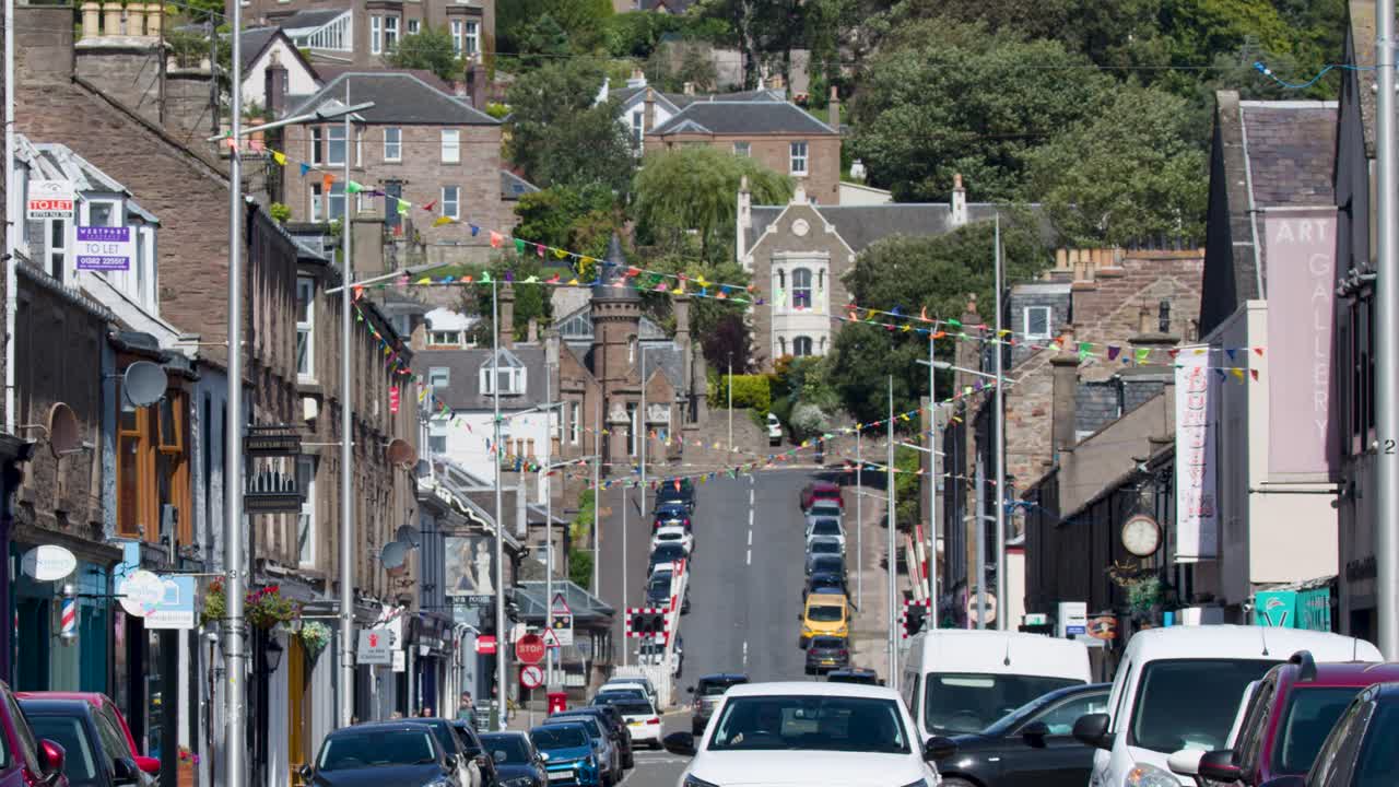 Cars drive uphill through Broughty Ferry town street, bright daylight, static wide shot