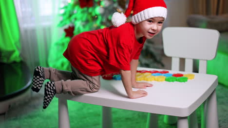 child plays with shapes by tree. a young child in a festive hat arranges colorful shapes on a table while surrounded by christmas decorations and a tree
