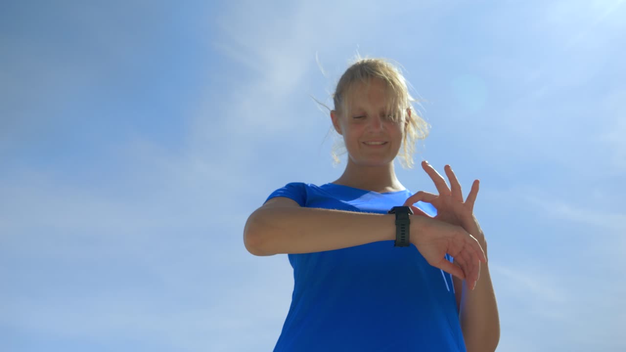 inquadratura dal basso di una donna bionda con una maglietta blu che usa il suo orologio collegato in una giornata di sole