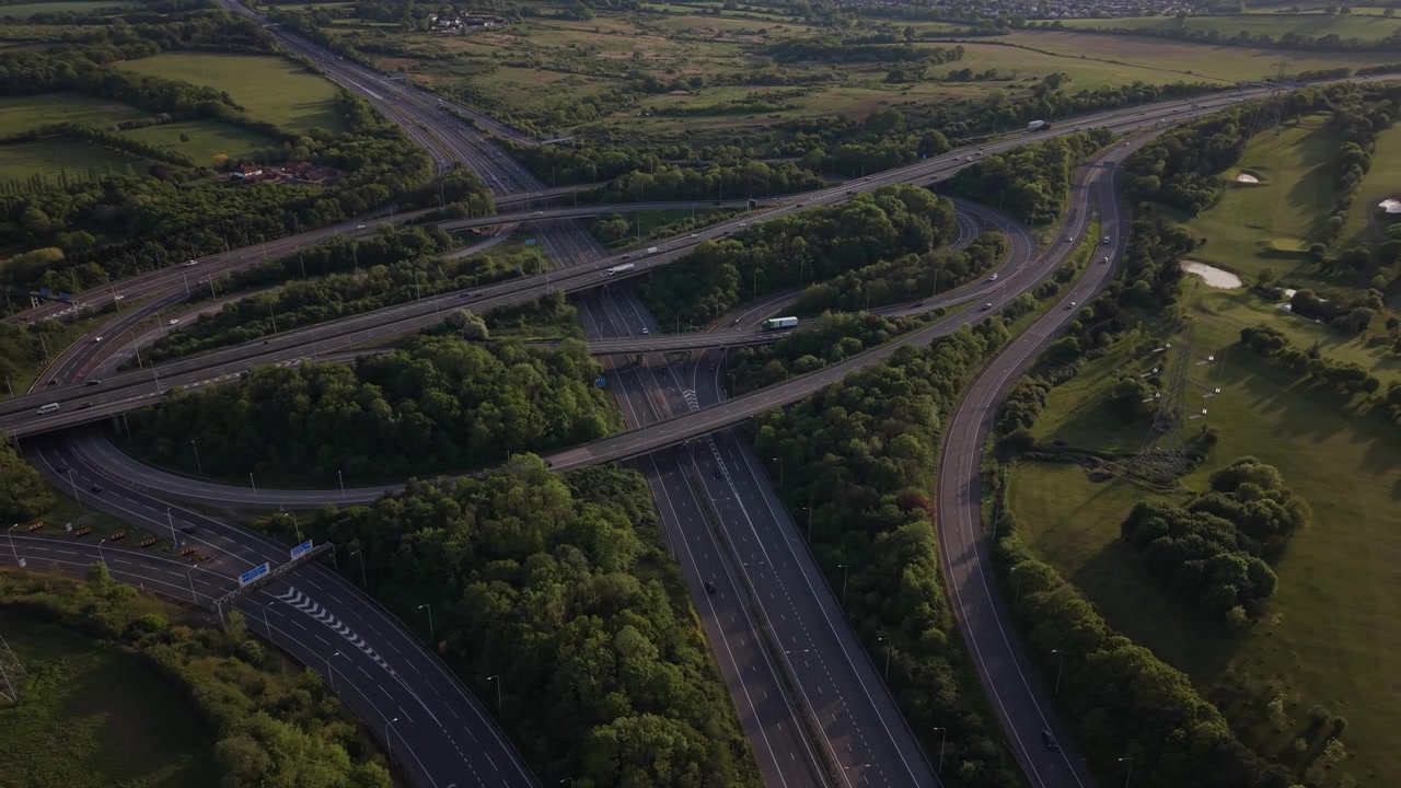 Aerial perspective shows a sprawling motorway interchange with vehicles navigating complex ramps. Green fields and trees surround the busy junction, bathed in soft late afternoon sunlight.
