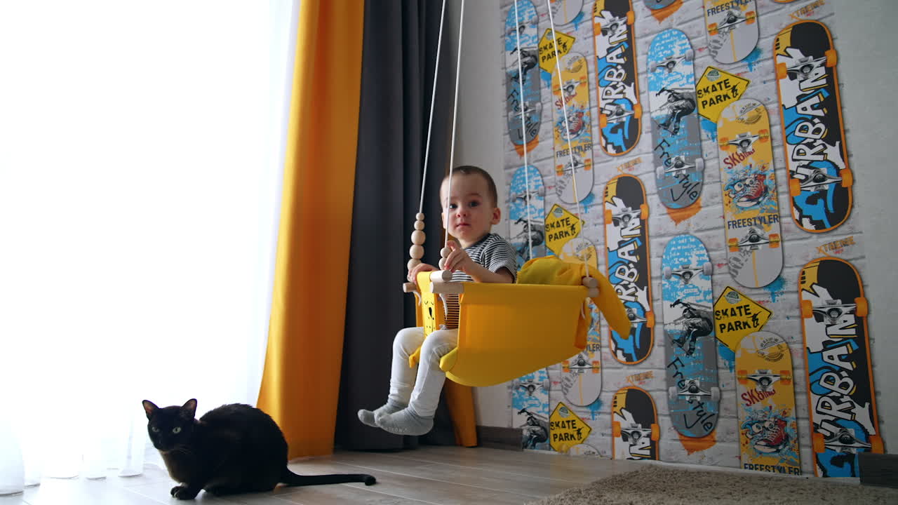Adorable baby boy swinging in the swing at home and smiles happily. Black cat sits on the floor beside. Low angle view.