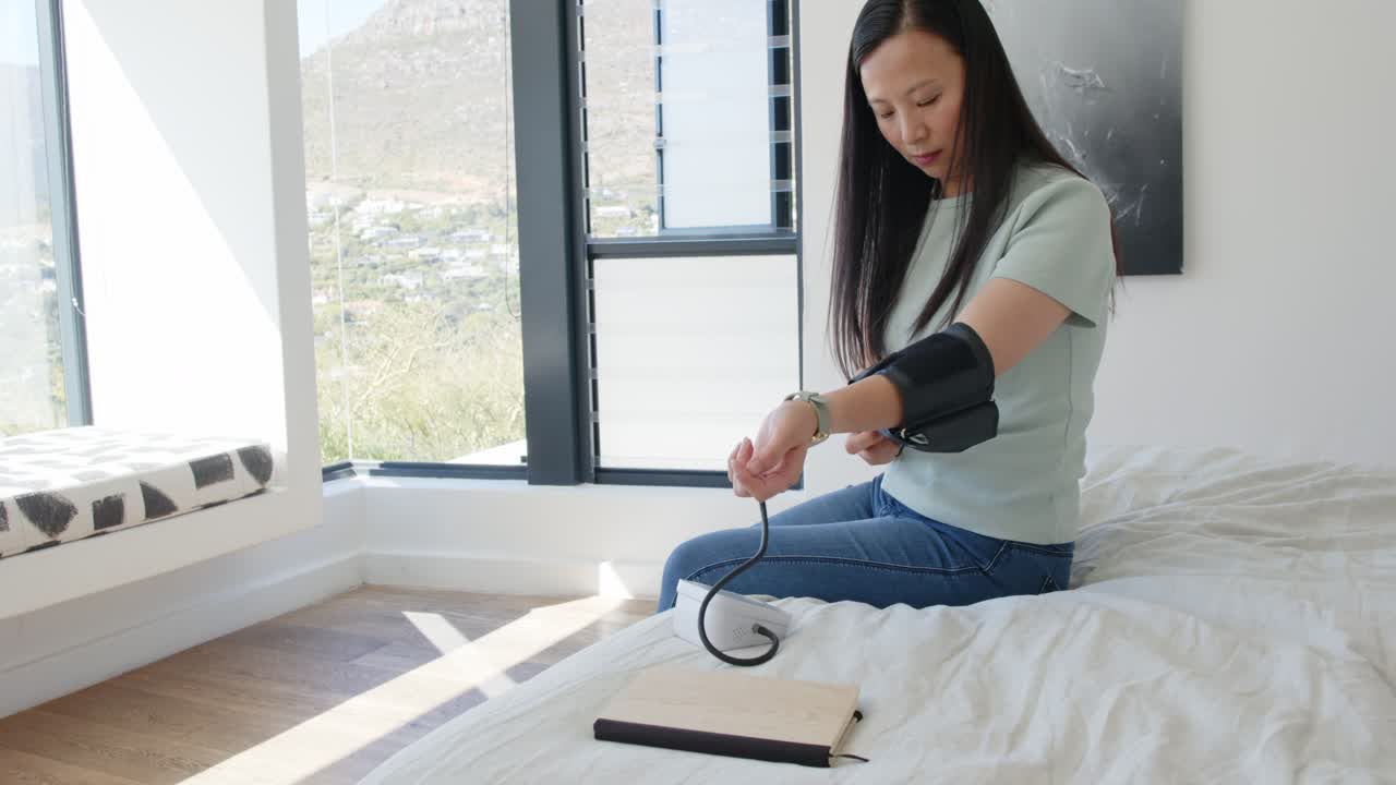 Donna asiatica a letto si allaccia il bracciale per la pressione sul braccio dopo aver controllato l'orologio da polso per misurare.