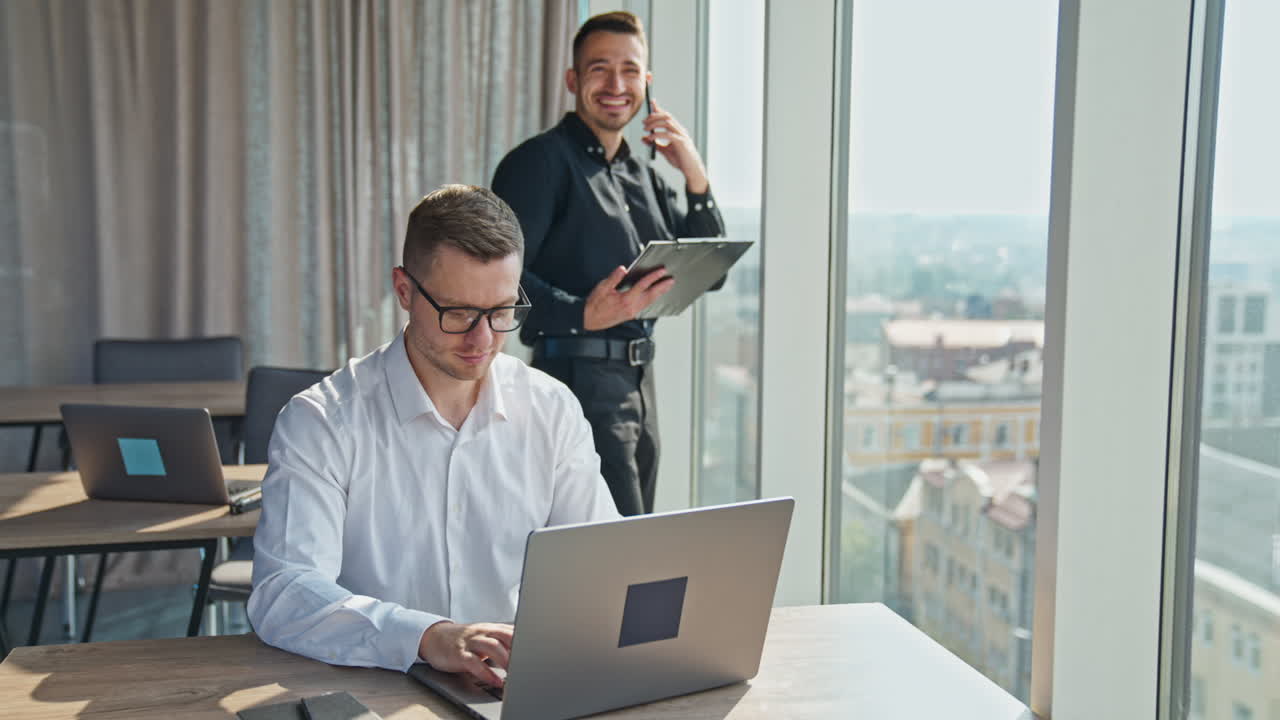 Young mid-aged office workers in the light spacious room. Male in white shirt works on laptop and man behind him talks on the phone and laughs.