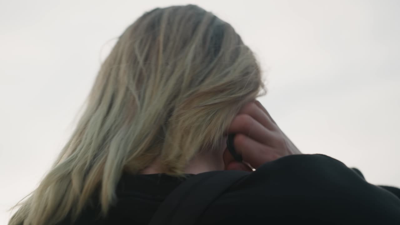 Mulher branca loira de costas, ao ar livre, amarrando o rabo de cavalo, com as mãos alisando as mechas. Cabelo com textura esvoaçada pelo vento, em um crepúsculo nublado. Ela veste um moletom preto com capuz, em um viaduto urbano, transmitindo um clima cinematográfico contemplativo, em um close-up íntimo de quietude e solidão.