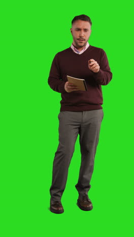 a man standing in front of a green screen, looking down at a notebook and pen in his hand.