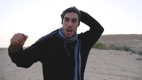 excited young man traveler looking at camera and screaming, shouting and shaking clenched fists raised up in the air while camping at crater ramon trail, negev desert, southern israel in slow motion