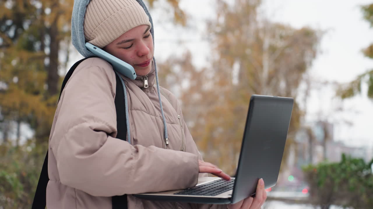 Close view of light skin lady in beige jacket and knitted hat multitasking outdoors in cold weather, typing on laptop while holding phone between ear and shoulder, with soft blurred autumn background