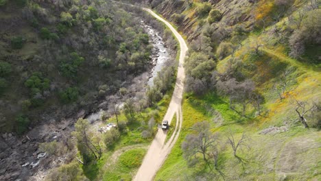 Drone-shot-of-a-black-pickup-truck-driving-along-a-river-with-wildflowers-in-the-mountains-on-a-dirt-road