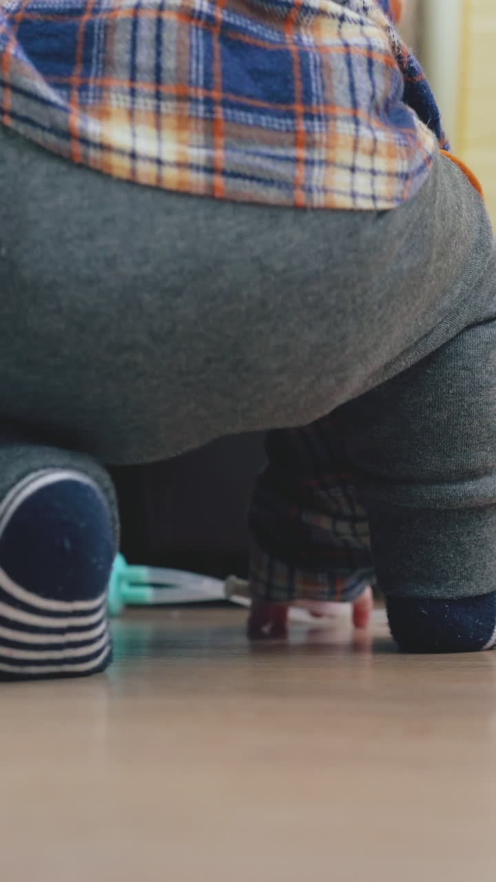 active baby in grey pants leans on soft bed standing on knees playing with aspirator on wooden floor close backside view