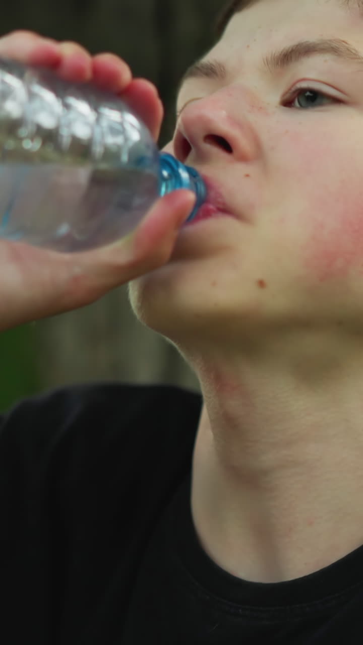 검은 티셔츠를 입은 어린 소년은 나무 에 앉아 플라스틱 병에서 물을 마시며, 따뜻한 날에 녹색으로 둘러싸인 신체 활동 후에 냉각하는 것처럼 보입니다.