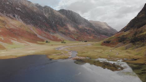 Abstieg-In-Ein-Schottisches-Hochlandtal,-Während-Die-Kamera-Entlang-Eines-Dunklen-Sees-Gleitet,-Der-Den-Bedeckten-Himmel-In-Glencoe,-Schottland,-Widerspiegelt