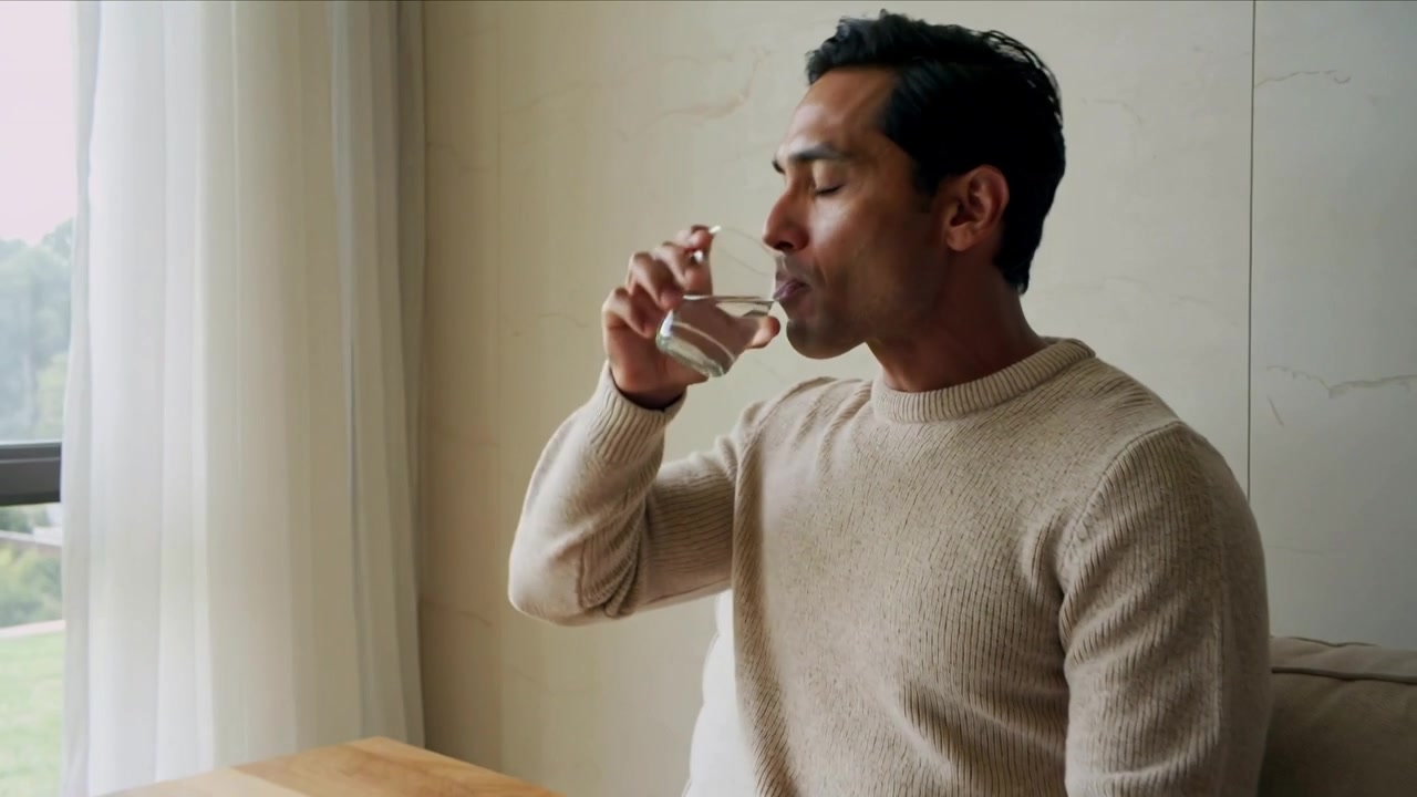 Drinking man in beige sweater hydrating placing glass on wooden table by sofa gazing out window