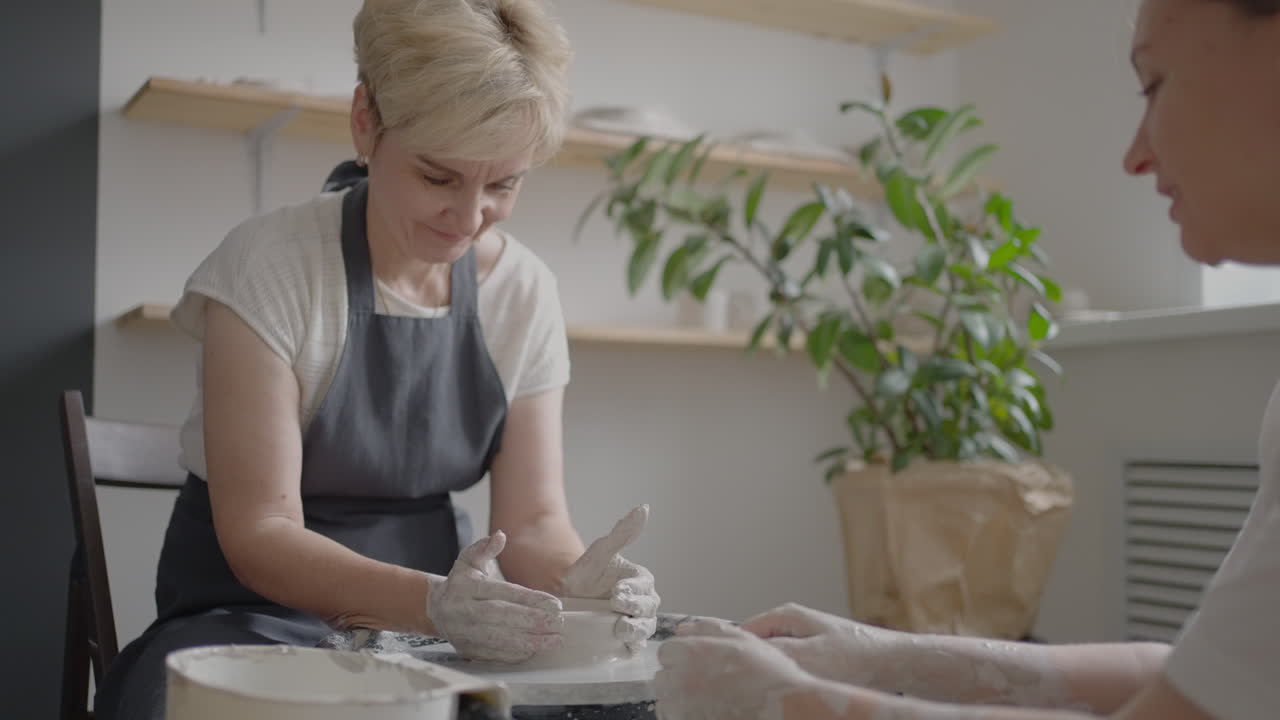 Woman master transfers knowledge to an elderly woman working on a potter's wheel and making a mug of ceramics in her workshop in slow motion