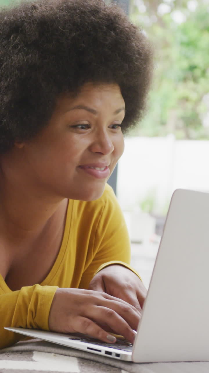 video de una feliz mujer afroamericana de tamaño grande acostada en el suelo y usando una computadora portátil