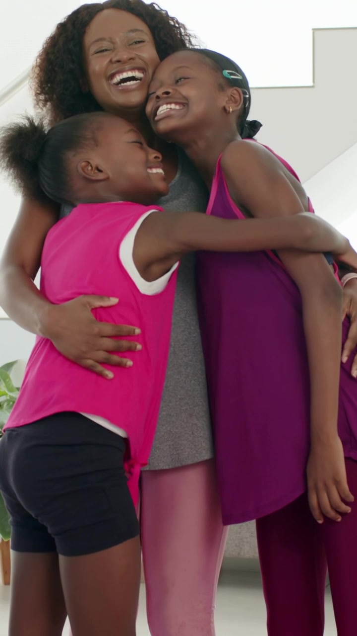 Vídeo vertical: Menina mais nova entrando pela esquerda, fazendo com que a mãe e as meninas se abracem perto do sofá, expressando alegria.